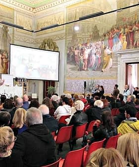 A large audience seated in a historical hall with elaborate murals on the walls. - Olive Oil Times
