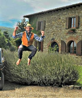 A man in a hat and vest jumping joyfully near a vintage car parked by a stone house. - Olive Oil Times