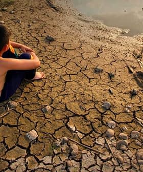 A child sitting on dry, cracked earth near a small body of water. - Olive Oil Times