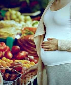 Pregnant woman standing in front of a fruit market display with various fruits. - Olive Oil Times
