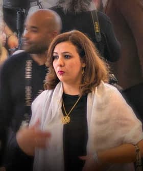 A woman wearing a white shawl and a necklace walking through a crowded social event. - Olive Oil Times