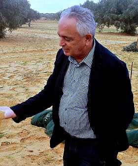 Man in a black jacket holding sand in his hand in an olive grove with a woman in the background. - Olive Oil Times