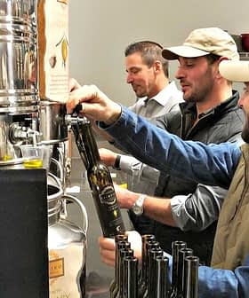 Three men filling bottles with olive oil from a large container in a processing facility. - Olive Oil Times