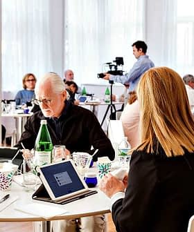 A group of individuals seated at tables during a meeting, with a photographer capturing the moment. - Olive Oil Times