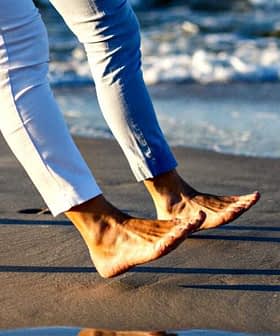 Two individuals walking barefoot along the shoreline with waves in the background. - Olive Oil Times