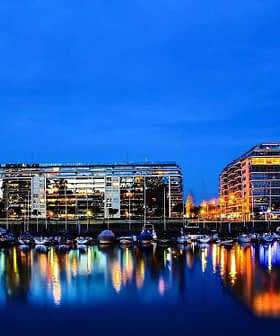 Night view of a city skyline with buildings and boats reflected in the water. - Olive Oil Times