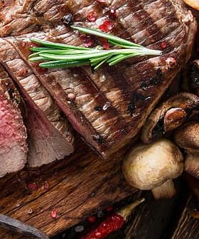 Sliced grilled steak on a wooden cutting board with tomatoes, mushrooms, and sauce. - Olive Oil Times