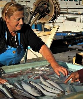 A woman in an apron selling fish at a market while a man points at the fish on display. - Olive Oil Times