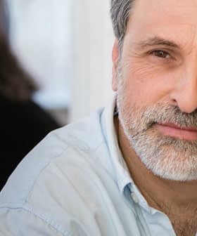 A man with a beard wearing a light blue shirt, engaged in conversation with another person. - Olive Oil Times