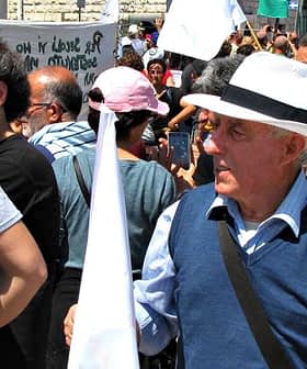 An older man wearing a white hat and holding a small flag at a crowded public event. - Olive Oil Times