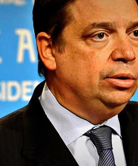 Man in a dark suit and tie speaking at a press conference with a blue background. - Olive Oil Times