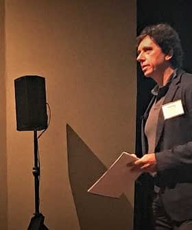 A man presenting at a conference with a microphone and projector screen in the background. - Olive Oil Times