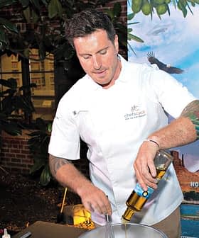Chef in a white uniform mixing ingredients in a bowl at an outdoor event with a backdrop. - Olive Oil Times