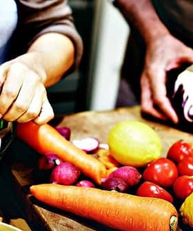 Hands preparing vegetables including lettuce, carrots, tomatoes, and cabbage on a wooden cutting board. - Olive Oil Times