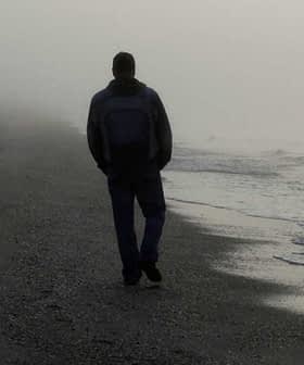 Individual walking alone on a foggy beach with gentle waves lapping at the shore. - Olive Oil Times