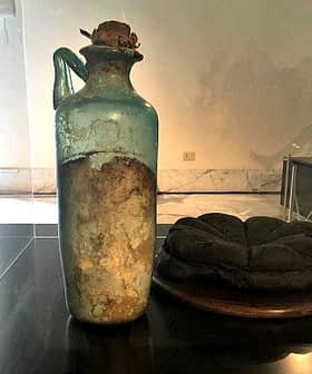 An ancient glass bottle with a cork stopper and a dark round object beside it on a display table. - Olive Oil Times