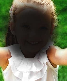 A child with outstretched arms smiling at the camera, wearing a white top with ruffles. - Olive Oil Times