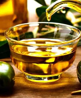 Glass container pouring olive oil into a small glass bowl with olives beside it. - Olive Oil Times