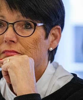 Woman with short black hair and glasses sitting at a table during a meeting, looking thoughtful. - Olive Oil Times