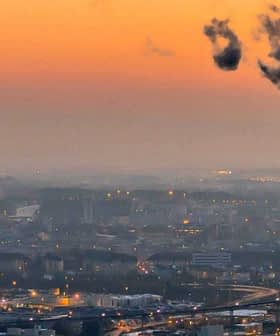 Smoke rising from a tall industrial chimney against a sunset sky with city skyline in the background. - Olive Oil Times