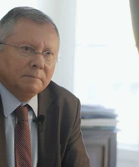 A man in a suit sitting at a desk with a serious expression, wearing glasses and a tie. - Olive Oil Times
