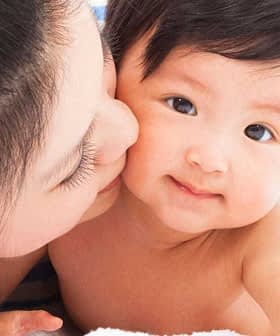 A mother kissing her baby on a soft surface, both looking at the camera. - Olive Oil Times