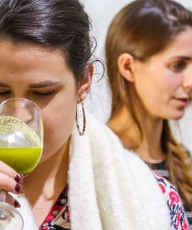 Woman holding a glass of green liquid while tasting olive oil at an event with two other women in the background. - Olive Oil Times