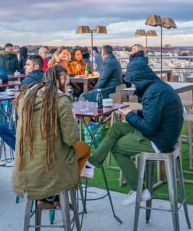 Group of people sitting and standing at a rooftop bar with tables and chairs. - Olive Oil Times