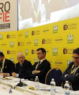 Panel of speakers at a conference on Agro-Mafie with a yellow backdrop and flags. - Olive Oil Times