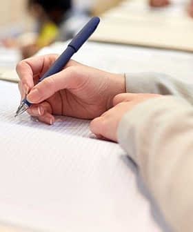 A person holding a pen and writing in a notebook during a classroom setting. - Olive Oil Times