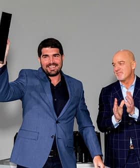 A man in a blue suit holding an award while two others applaud in the background. - Olive Oil Times