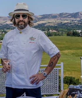 Chef wearing a white coat, hat, and sunglasses standing outdoors with a scenic background. - Olive Oil Times
