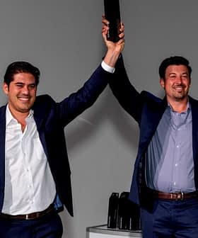 Two men in suits raising a trophy above their heads in a celebratory manner. - Olive Oil Times