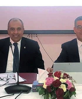 Four individuals seated at a conference table with a flag and floral arrangement in front of them. - Olive Oil Times