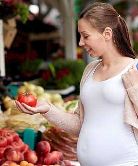 Pregnant woman examining a tomato while shopping at a farmers market with various fruits and vegetables around her. - Olive Oil Times