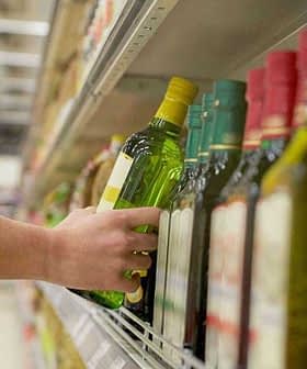Individual reaching for a green bottle of olive oil on a supermarket shelf among other oil bottles. - Olive Oil Times