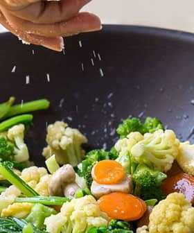 Person sprinkling salt over a mix of vegetables in a black wok with a wooden spoon. - Olive Oil Times