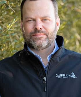 Man with a beard wearing a black vest featuring the California Olive Ranch logo. - Olive Oil Times