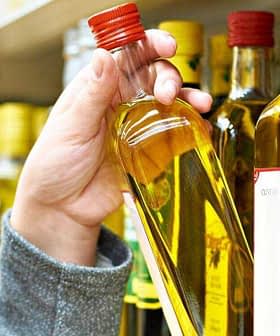 A person holding a bottle of olive oil from a shelf in a grocery store. - Olive Oil Times