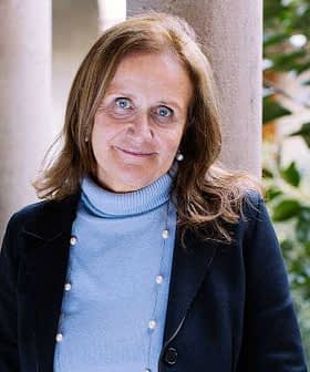 Woman with brown hair wearing a blue sweater and black jacket standing in a corridor with columns. - Olive Oil Times