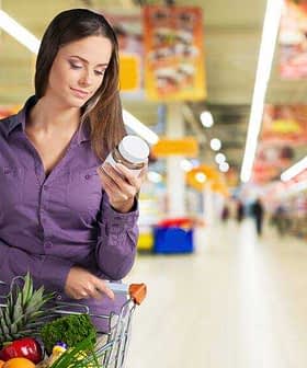 Woman examining a product label while holding a jar in a grocery store aisle. - Olive Oil Times