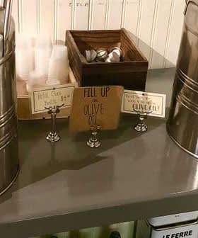 Two stainless steel dispensers labeled for olive oil and vinegar on a table with utensils. - Olive Oil Times