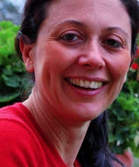 A woman smiling while holding a pot of flowers with red blooms outdoors. - Olive Oil Times