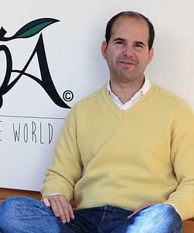 Man wearing a yellow sweater sitting on the floor in front of the LOA logo. - Olive Oil Times