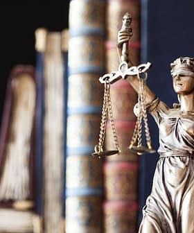 Bronze statue of Lady Justice holding scales and wearing a blindfold, with books in the background. - Olive Oil Times