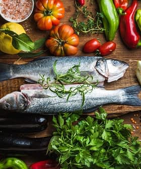 Two whole fish placed on a wooden board surrounded by various fresh vegetables and herbs. - Olive Oil Times