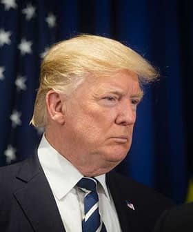 Man with blonde hair wearing a suit and tie, facing to the side against a dark background with flags. - Olive Oil Times