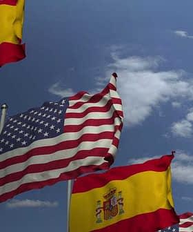 Spanish flags and an American flag waving in the wind against a blue sky with clouds. - Olive Oil Times