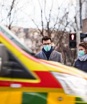 Two individuals wearing masks waiting at a crosswalk as an ambulance drives by in the foreground. - Olive Oil Times