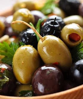 A wooden bowl filled with a variety of green and black olives, garnished with herbs. - Olive Oil Times